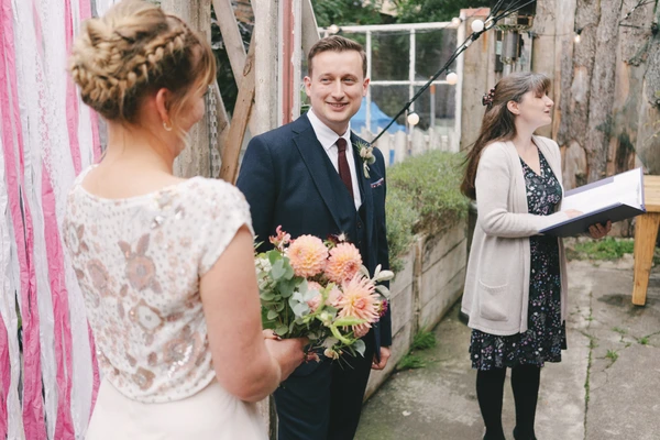 Bride in lace with orange bouquet looks at the smiling groom — female officiant beside them