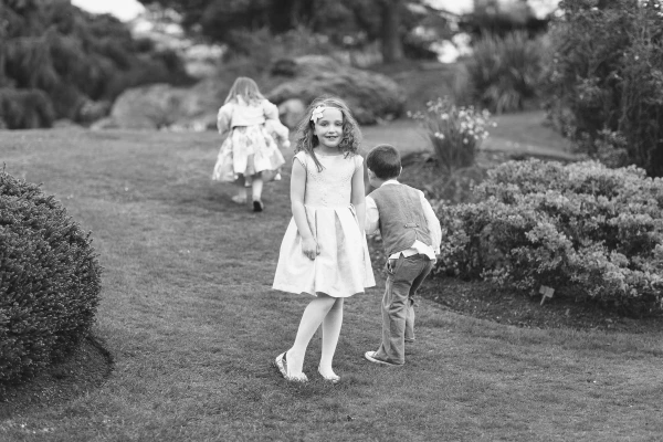 Black and white image of a flower girl pausing on the garden lawn and looking back at the camera with a wide smile while another child runs in the background. Candid documentary wedding photography.