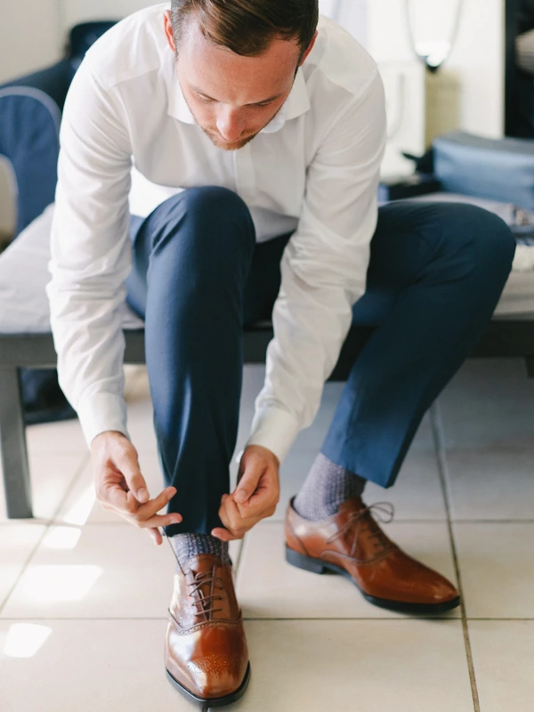 Groom in white shirt and blue trousers crouches to tie his brown shoes — focused, bright room