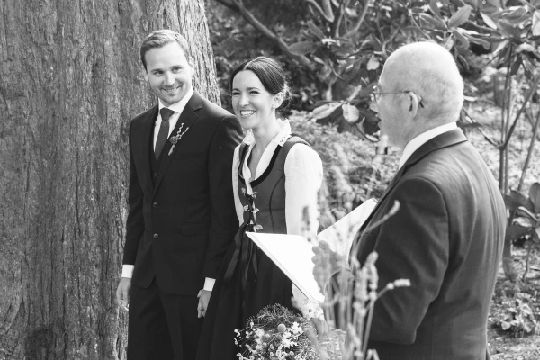 Black and white candid shot of the couple smiling together during the outdoor ceremony next to a large tree trunk, guests visible in the background. Intimate ceremony moment.
