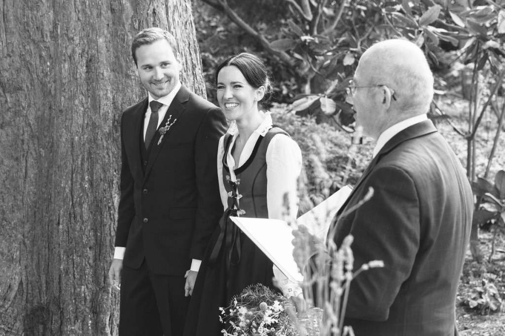 Black and white candid shot of the couple smiling together during the outdoor ceremony next to a large tree trunk, guests visible in the background. Intimate ceremony moment.