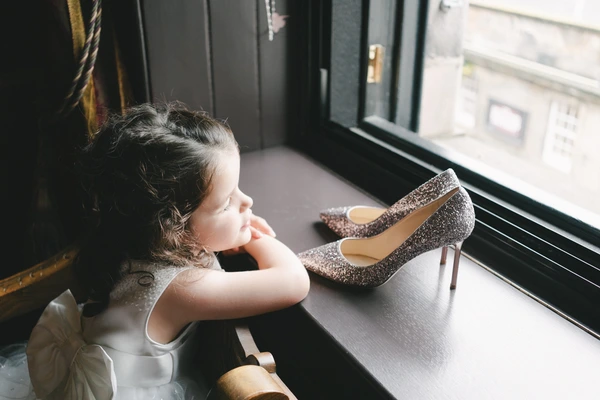 Little girl looks thoughtfully at the window, silver bridal shoes on the table beside her