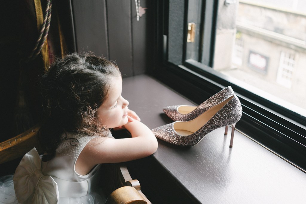 Little girl looks thoughtfully at the window, silver bridal shoes on the table beside her