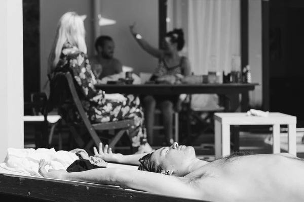 Boy lying peacefully on a bed in foreground, long-haired woman and others chatting in the room