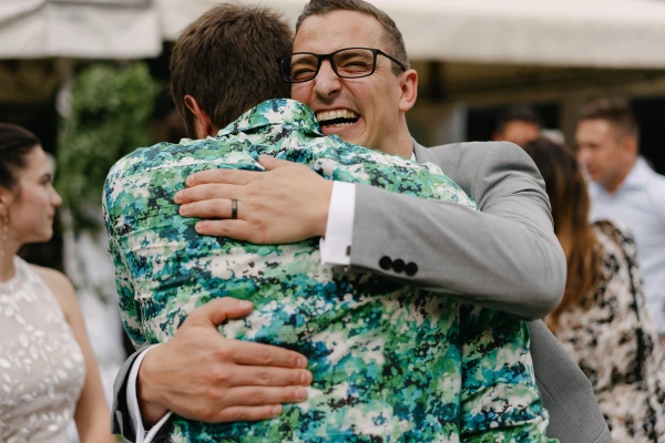 Groom hugging guest in green floral shirt at outdoor wedding reception with happy expression.