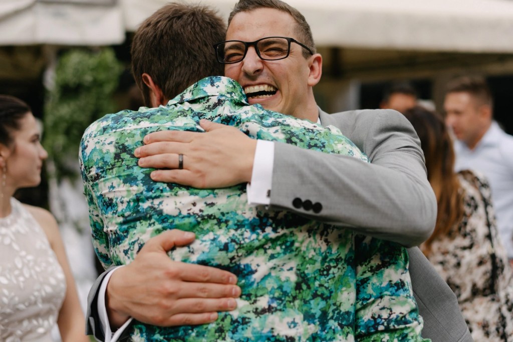 Groom hugging guest in green floral shirt at outdoor wedding reception with happy expression.
