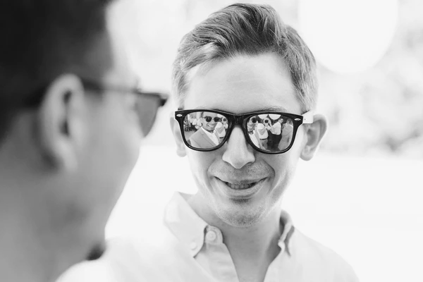 Boy in black-framed sunglasses smiling — mirror lenses reflect the wedding guests, white shirt