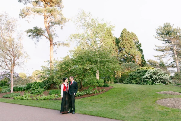 A wide shot of the couple standing together on a garden path in the Royal Botanic Garden Edinburgh surrounded by lush trees and spring flowers in the evening light. Documentary wedding portrait.