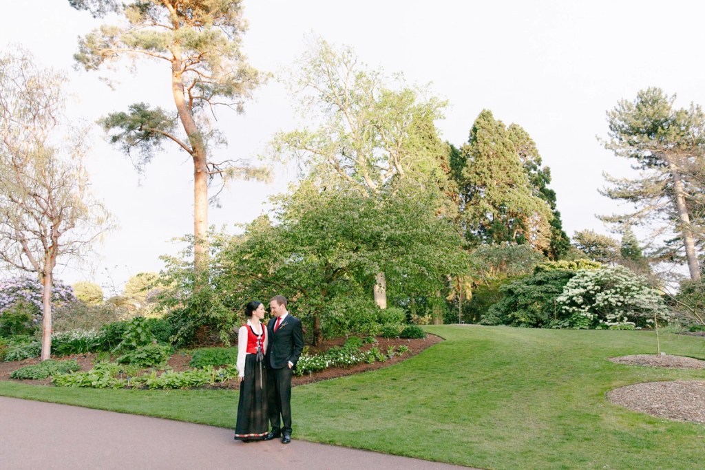 A wide shot of the couple standing together on a garden path in the Royal Botanic Garden Edinburgh surrounded by lush trees and spring flowers in the evening light. Documentary wedding portrait.