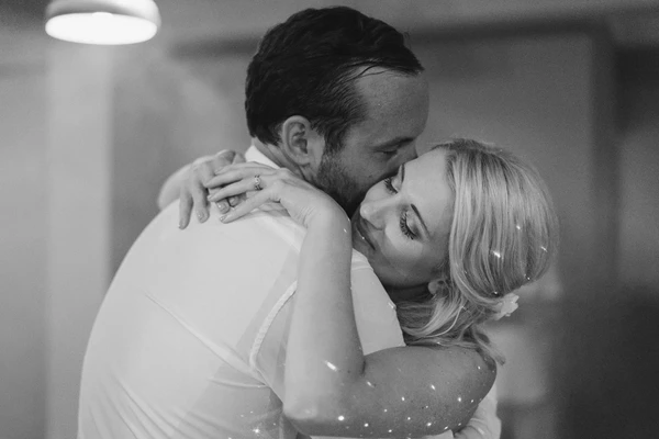 Groom kisses bride after their first dance — white sparkling gown, pearl hair pins, warm spotlight