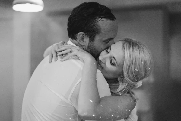 Groom kisses bride after their first dance — white sparkling gown, pearl hair pins, warm spotlight