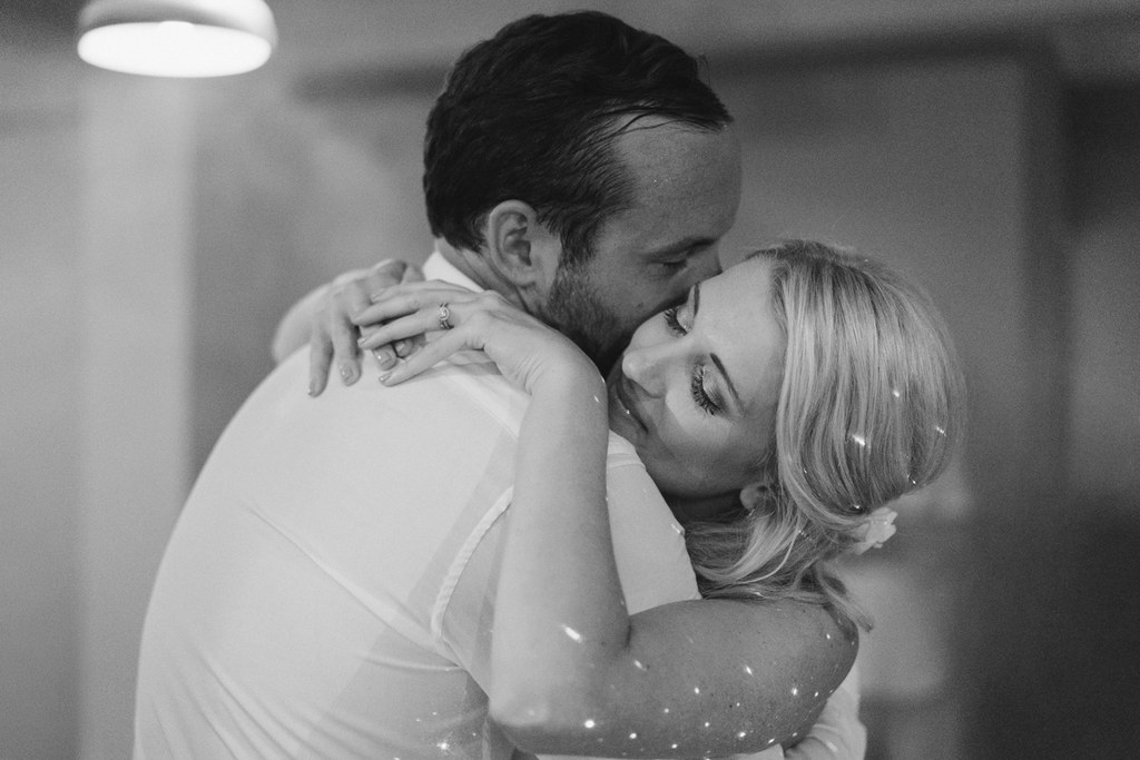 Groom kisses bride after their first dance — white sparkling gown, pearl hair pins, warm spotlight