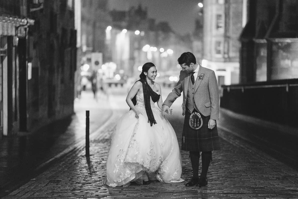Bride and groom strolling at night — white lace, black stole, grey suit with tartan. Black-and-white.