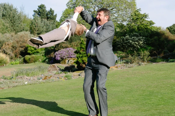 A man in a suit swings a young child through the air on a garden lawn, laughing with open delight. Candid documentary wedding photography.