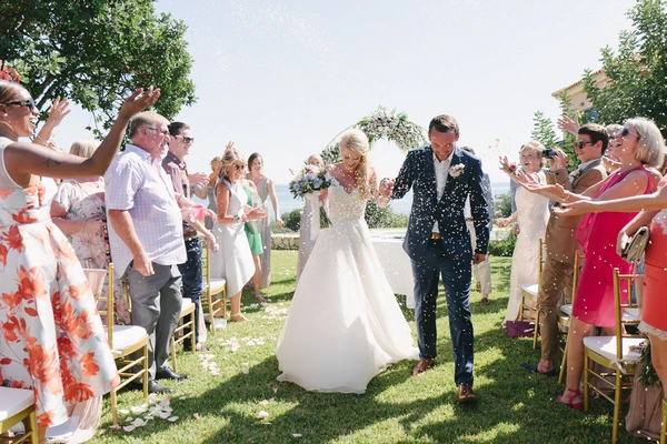 Young couple walks the ceremony aisle as guests cheer and toss flowers — Tuscan villa behind