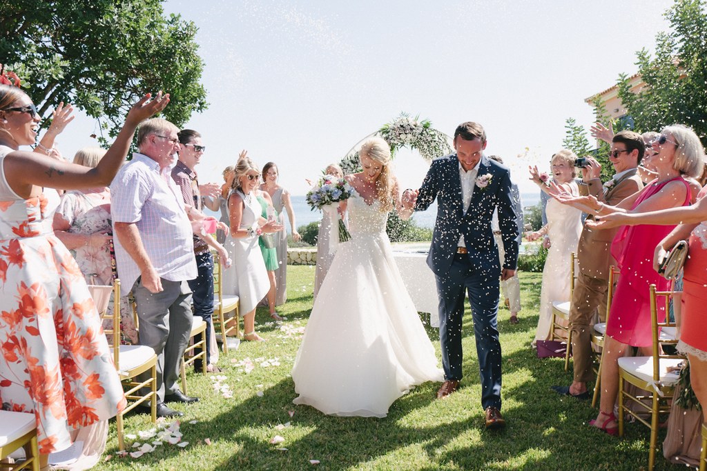 Young couple walks the ceremony aisle as guests cheer and toss flowers — Tuscan villa behind