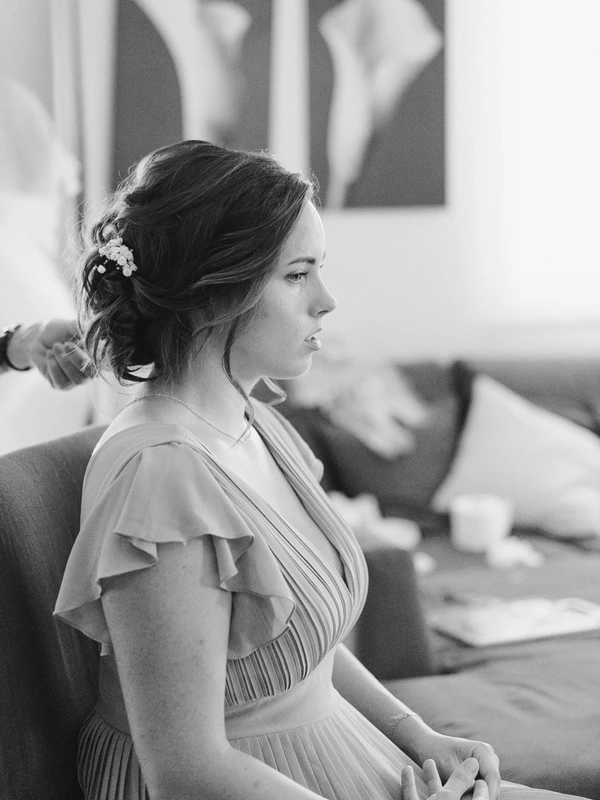 Bride in profile with white floral hair detail, pleated dress — pensive, natural light. Black-and-white.