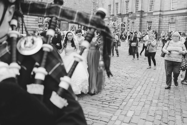 Bride with long white veil and floral crown before a stone courtyard, guests and officials around