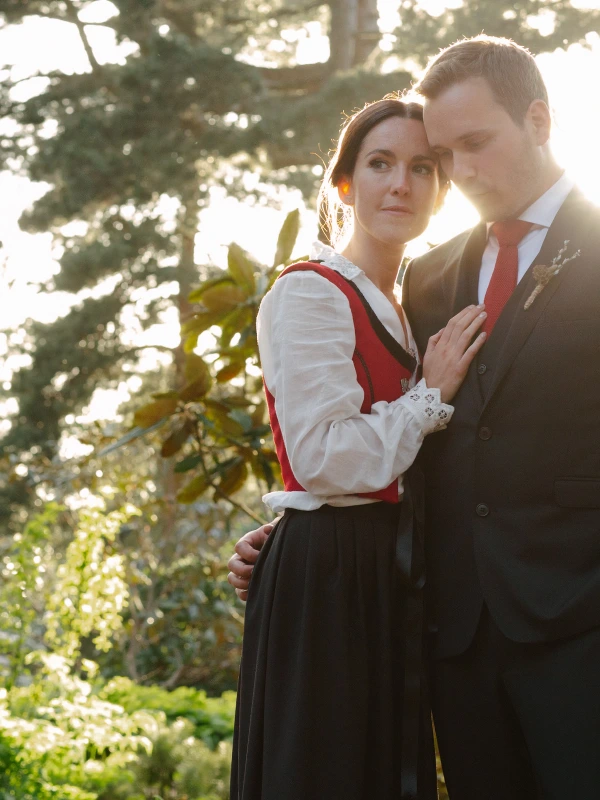 The bride in a Norwegian bunad rests her head against the groom's cheek as golden evening light streams through trees behind them. Intimate documentary wedding portrait at the Royal Botanic Garden Edinburgh.
