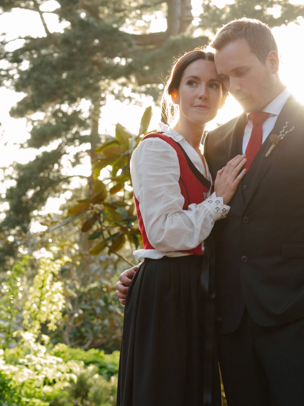 The bride in a Norwegian bunad rests her head against the groom's cheek as golden evening light streams through trees behind them. Intimate documentary wedding portrait at the Royal Botanic Garden Edinburgh.