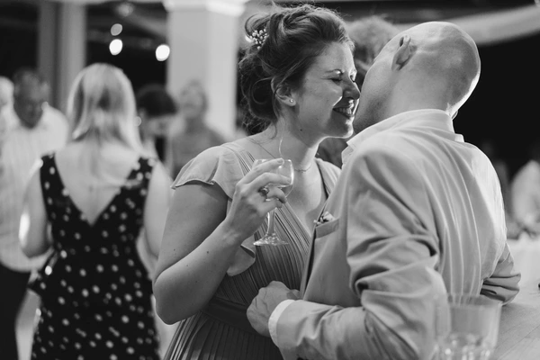 Bride and groom dance at the reception, she smiles up at him — guest in polka-dot dress behind. B&W.