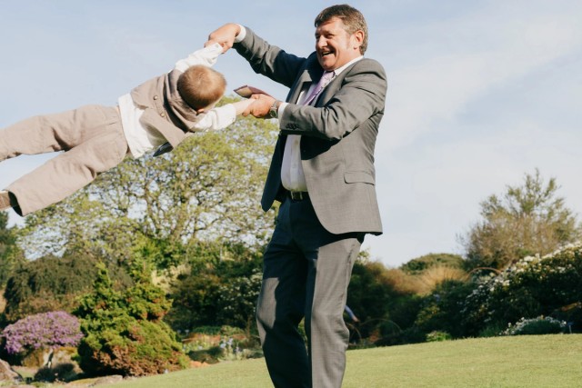 Man in gray suit swinging laughing child through air in sunny garden during wedding celebration