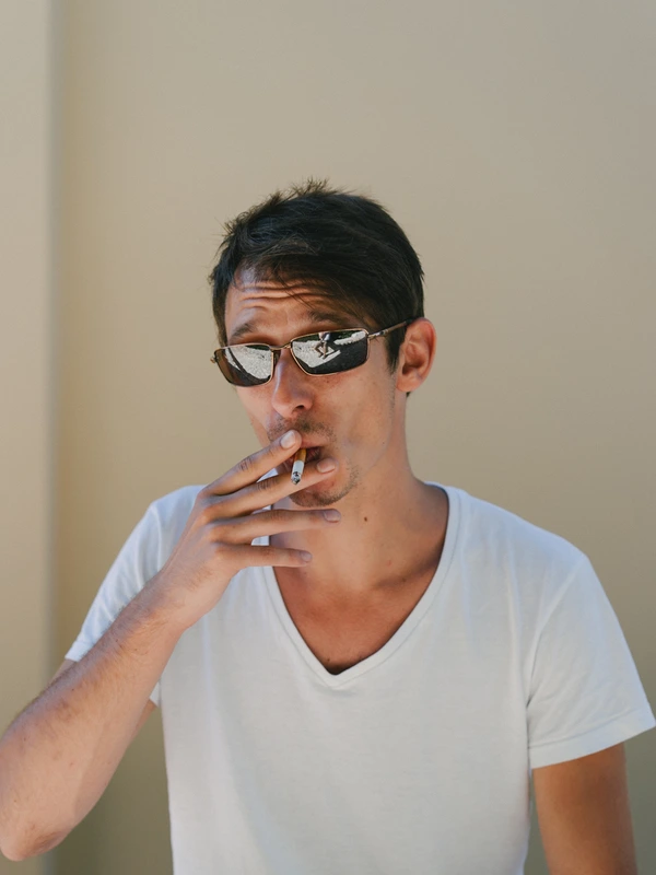 Boy in sunglasses and white polo with a cigarette, thoughtful look, leaning against a plain wall