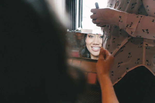 Bride smiles naturally in the mirror as a helping hand adjusts her white hat — joy and anticipation