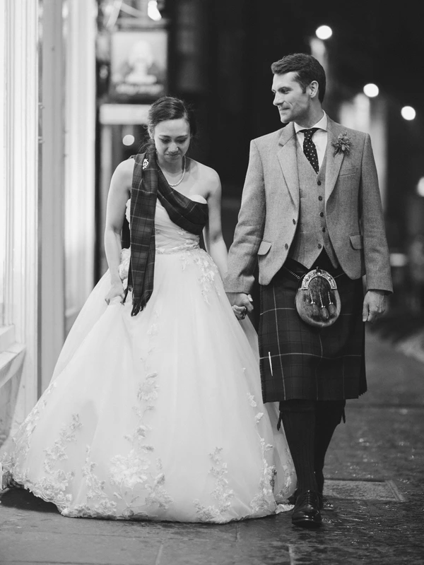 Newlyweds hand in hand on a night street — groom in tartan, bride in white lace, shadow and light