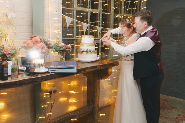 Bride and groom cut the wedding cake together — pink flowers and warm fairy lights