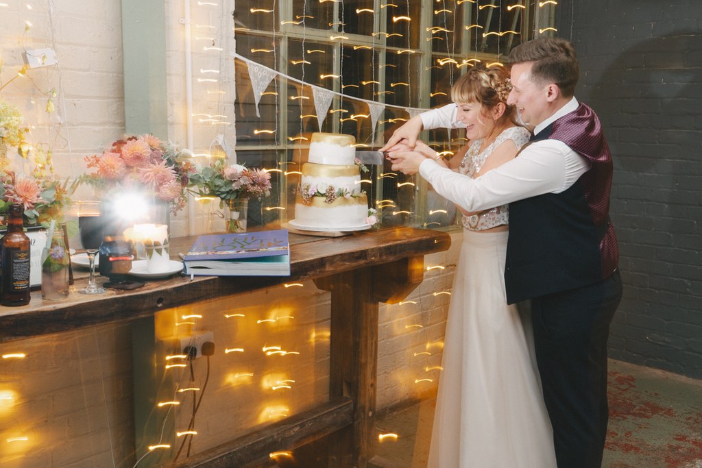 Bride and groom cut the wedding cake together — pink flowers and warm fairy lights