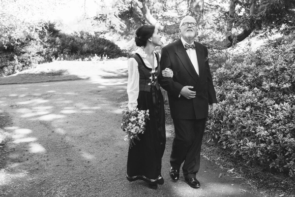 Black and white candid shot of the bride in her Norwegian bunad walking arm in arm with her father through the Royal Botanic Garden Edinburgh before the ceremony. Tender pre-ceremony moment.