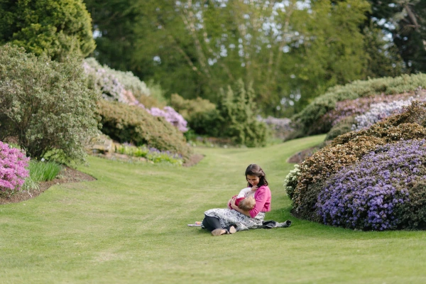 A woman in a bright pink top sits on the grass cradling a baby among flowering shrubs in the Royal Botanic Garden Edinburgh. Candid documentary wedding moment.