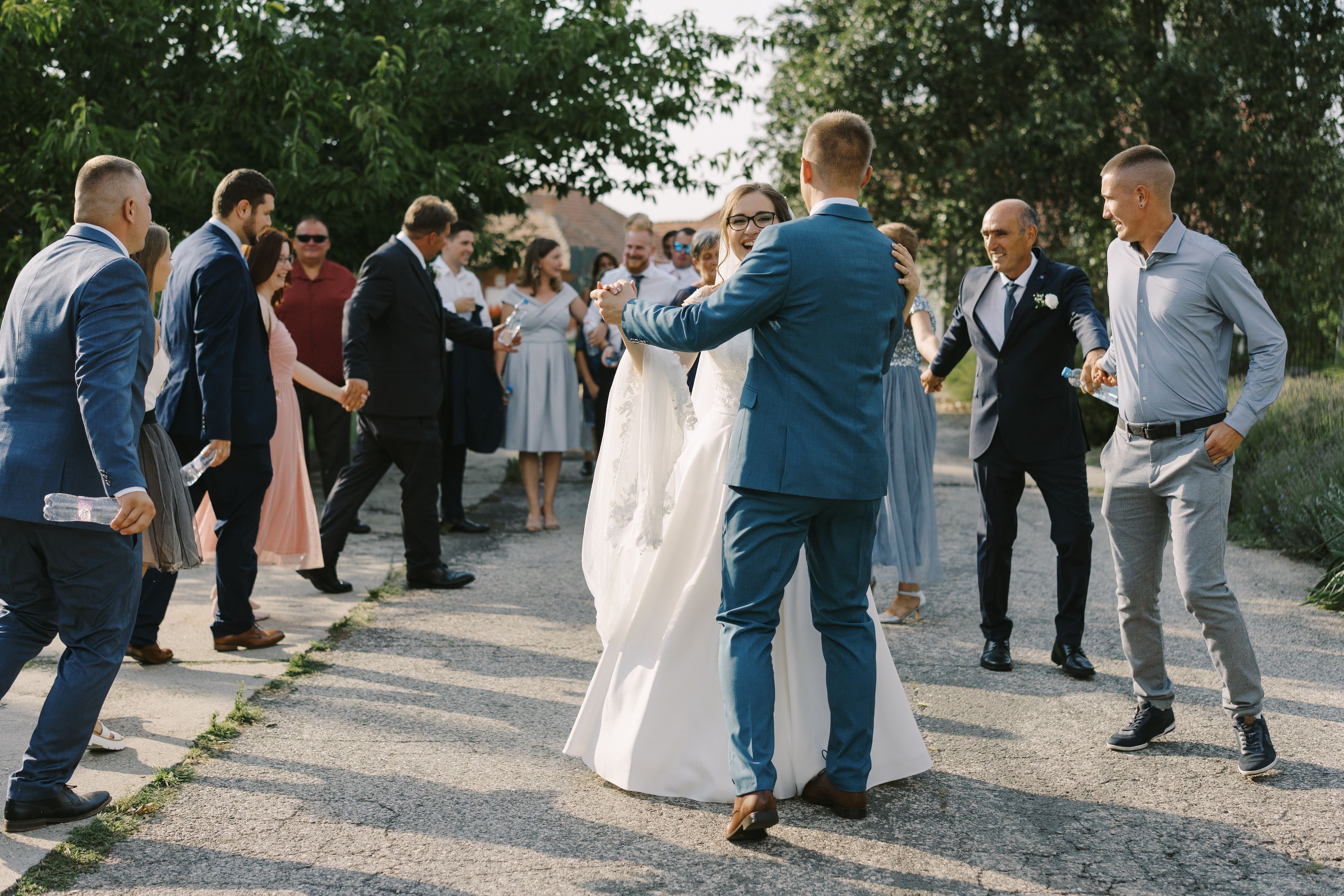 Vőlegény fehér ruhában lévő menyasszonnyal táncol, vendégek körülötte egy kertben, vidám esküvői ünneplés a természetben.