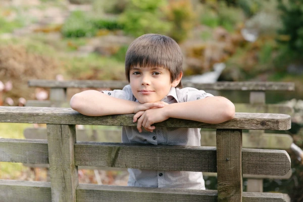 A young boy leans his chin on his folded arms on a wooden fence railing, looking thoughtfully into the camera. Candid portrait from a wedding at the Royal Botanic Garden Edinburgh.