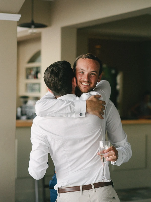 Groom in white shirt smiles receiving a friend's hug, champagne glass in hand, modern kitchen