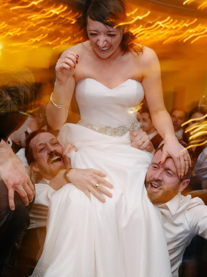 Bride in white gown laughing joyfully as two men lift her on their shoulders during lively wedding reception dance floor celebration.