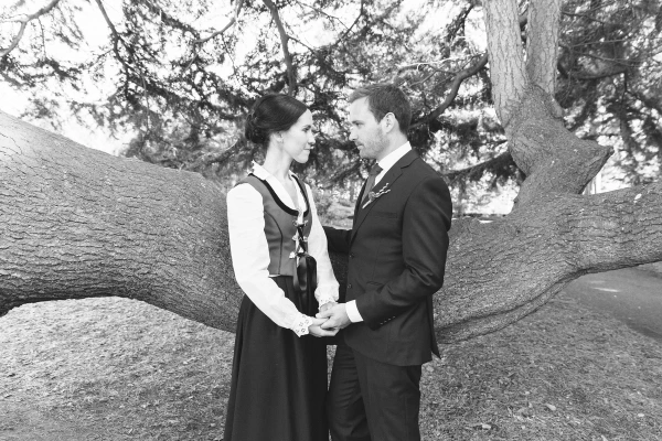 Black and white portrait of the couple holding hands and looking at each other tenderly while leaning against a large fallen tree trunk in the Royal Botanic Garden Edinburgh. Romantic documentary portrait.