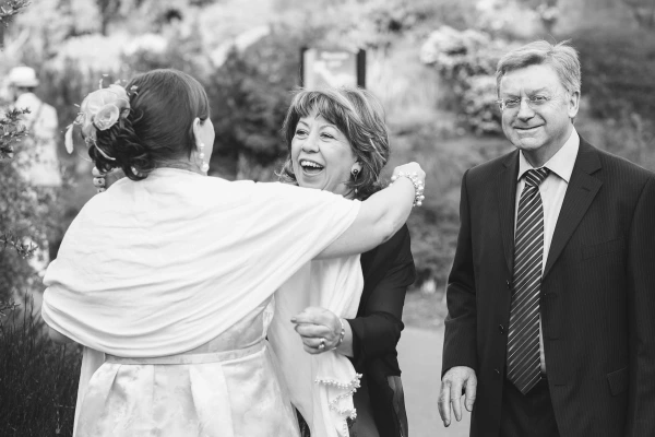 Black and white image of the bride in a white wrap embracing a laughing woman, while a man in a suit and glasses watches warmly in the garden. Candid documentary wedding photography.