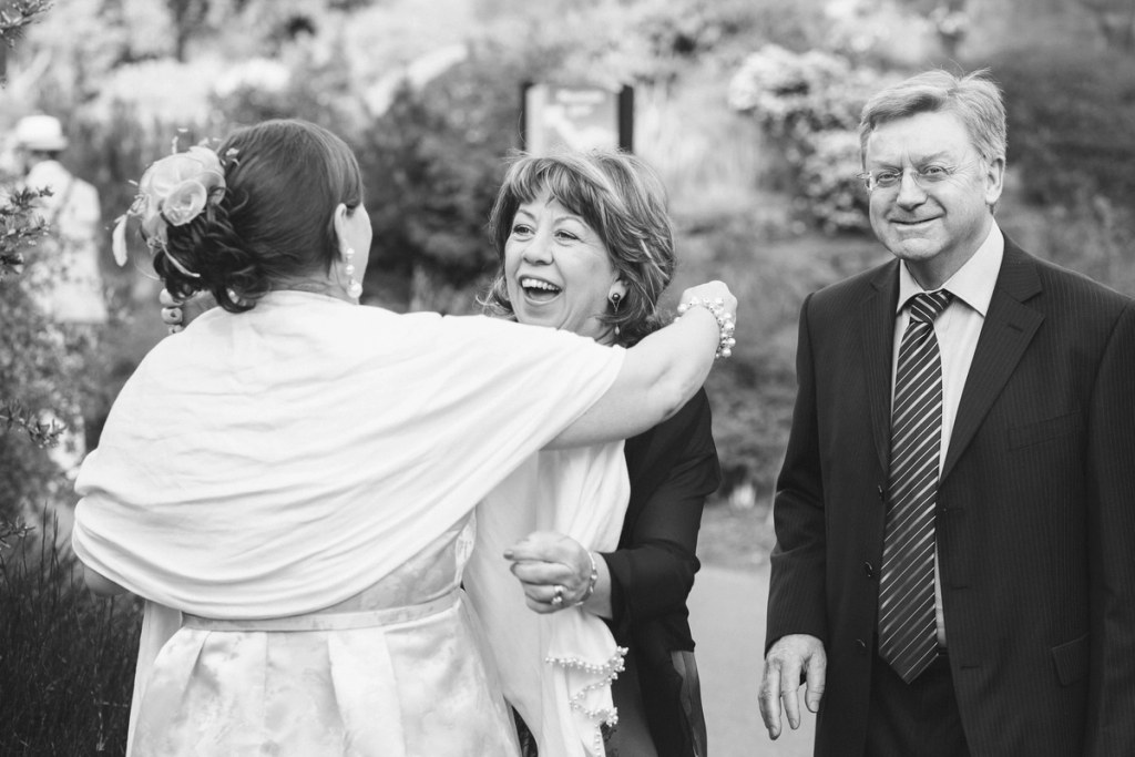 Black and white image of the bride in a white wrap embracing a laughing woman, while a man in a suit and glasses watches warmly in the garden. Candid documentary wedding photography.