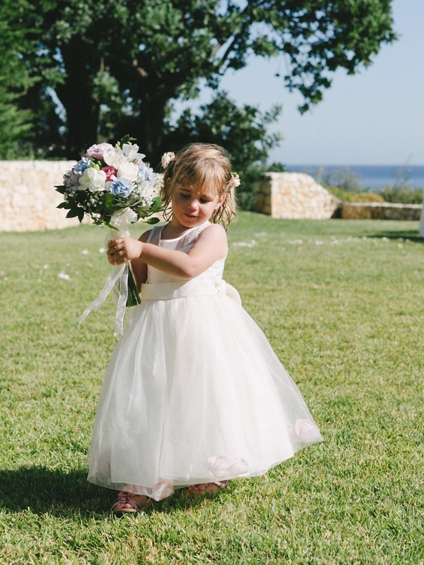 Little girl in white tulle with bouquet stands smiling on green lawn — large trees and sea behind