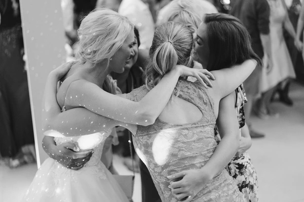 Bride in sparkling white gown hugs friends in a loving moment — black-and-white