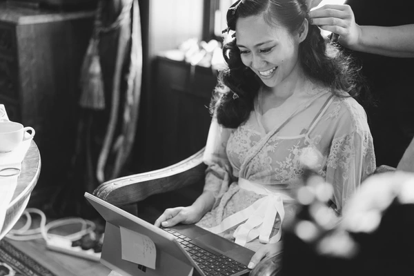 Smiling bride at the laptop during hair styling — natural light, black-and-white