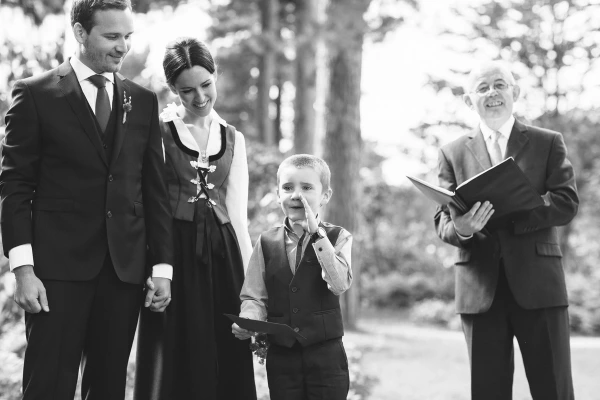 Black and white candid shot of the couple holding hands during the ceremony as a young boy standing beside them adorably picks his nose, the officiant smiling behind. Charming documentary moment.