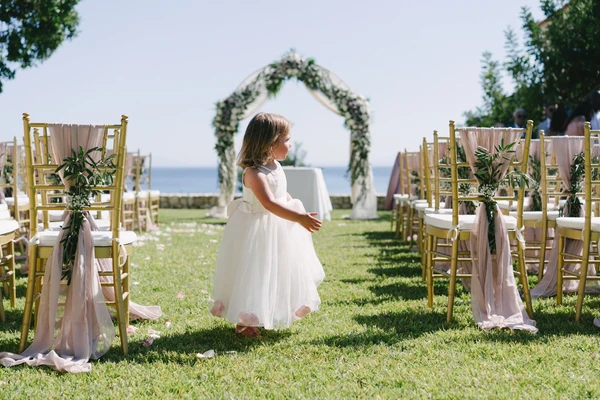 Little girl in white stands at the ceremony venue among gold chairs and pink ribbons — floral arch