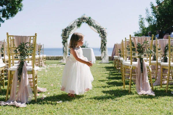 Little girl in white stands at the ceremony venue among gold chairs and pink ribbons — floral arch