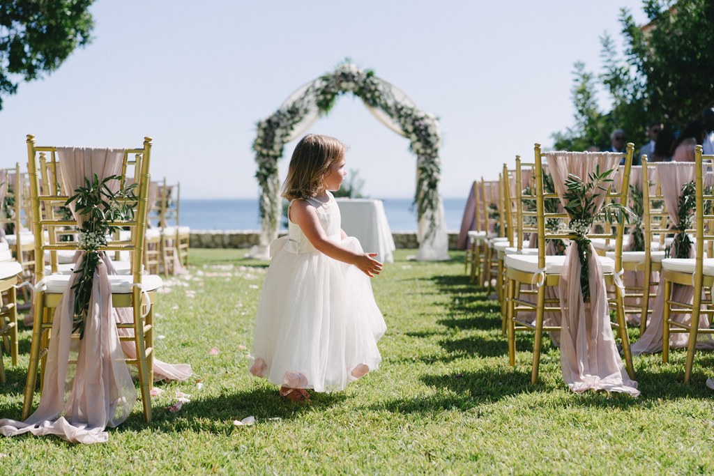 Little girl in white stands at the ceremony venue among gold chairs and pink ribbons — floral arch