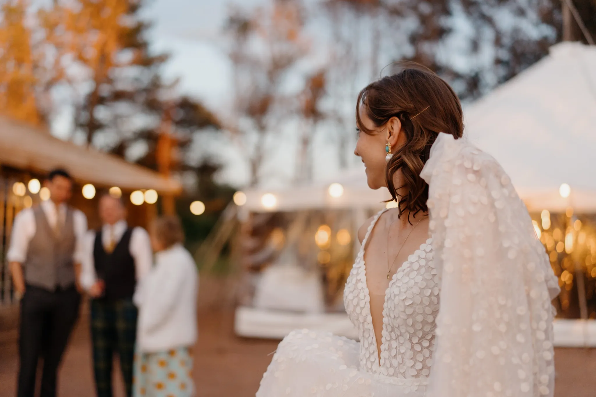 Bride in textured white gown and veil smiles outdoors at twilight wedding venue with string lights and guests in background.