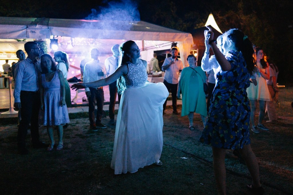 Wedding guests dancing outdoors at night celebration with bonfire and atmospheric lighting.