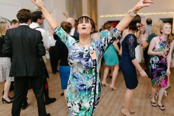 The bride in her Norwegian bunad laughs with a red-bearded man at the wedding reception, her hand gently touching his face. Warm candid documentary wedding photograph.